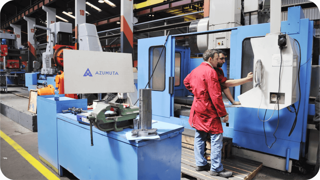 Two workers operate a large machine in an industrial factory setting. One in a red coat looks at the screen, while the other adjusts the controls. Nearby, various machinery and tools, including a vise, are visible. An 