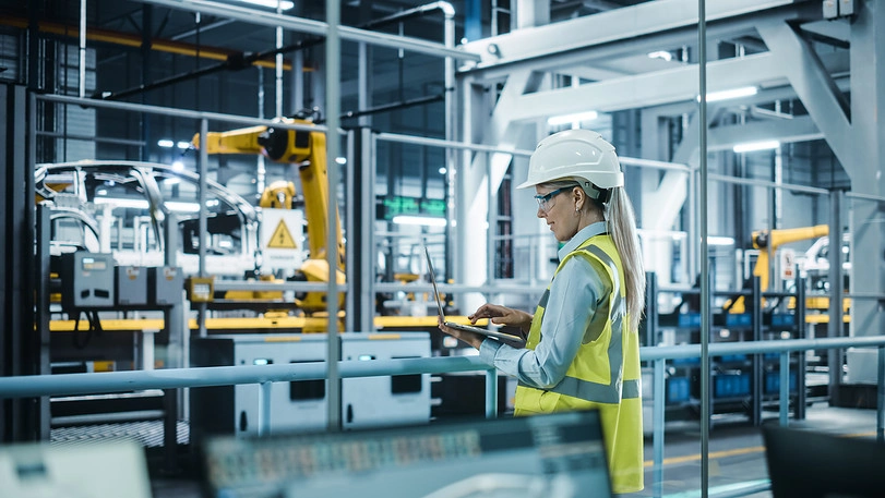 A woman wearing a hard hat and safety vest operates a tablet in a modern automobile factory. Robotic arms and machinery are visible in the background, indicating an advanced, automated production environment. She appears to be inspecting or monitoring the process.