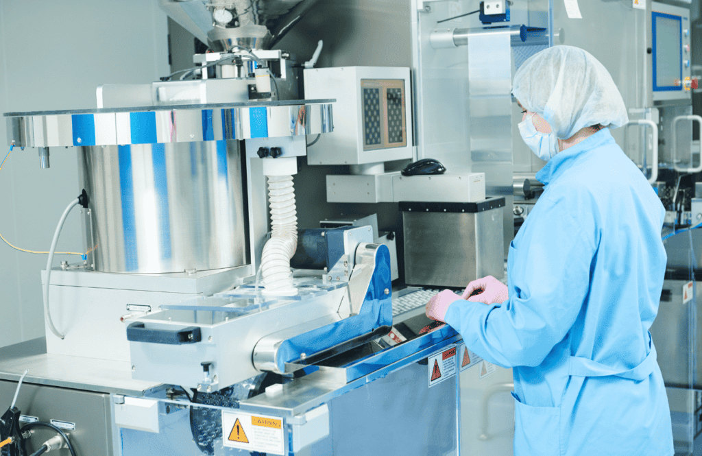 A lab technician in protective attire, including a hairnet, face mask, and gloves, operates large, stainless steel machinery in a cleanroom environment. The lab is equipped with advanced techno, and there is a screen displaying data on one of the machines.