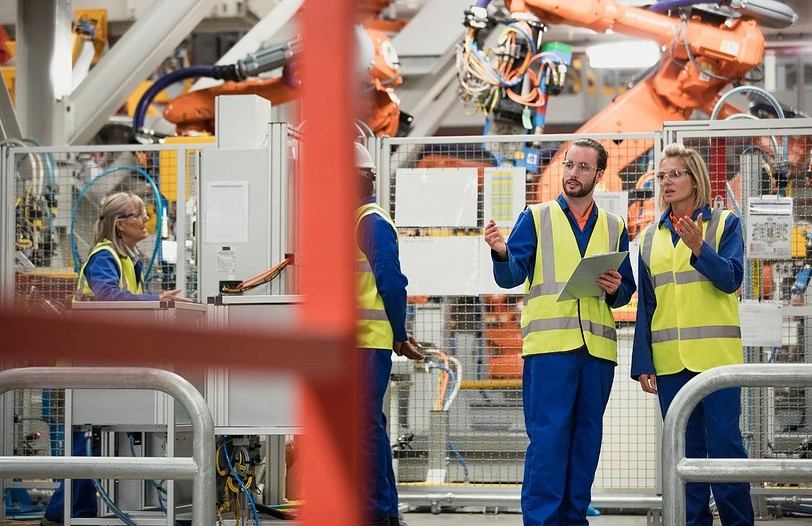 Four workers in a factory, wearing blue uniforms and yellow safety vests, engage in discussion. The setting features machinery and industrial robots in the background, indicating an active manufacturing environment.