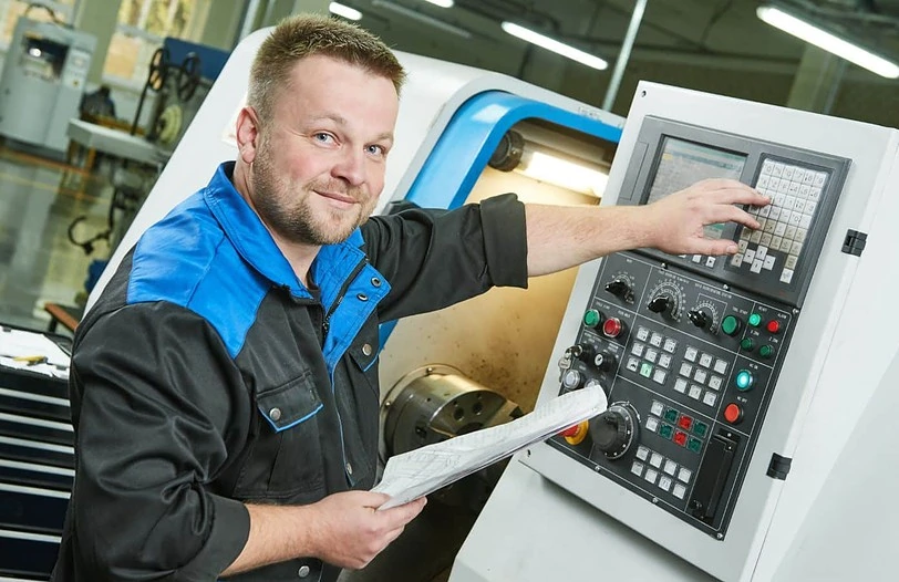 A man with a beard and short hair operates a CNC machine. He is wearing a black and blue work jacket and is holding a piece of paper in his left hand while pressing buttons on the machine's control panel with his right hand. He is smiling at the camera.