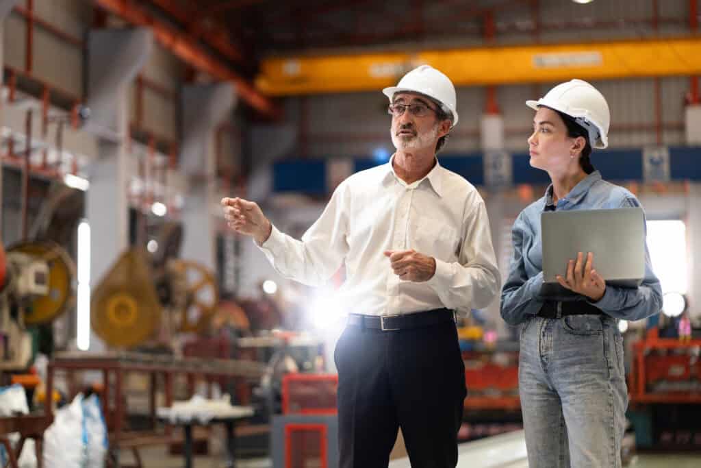 Two engineers wearing white hard hats stand in a manufacturing facility. The older engineer in a white shirt gestures while talking, and the younger engineer in a blue shirt holds a laptop. Industrial equipment and machinery are visible in the background.