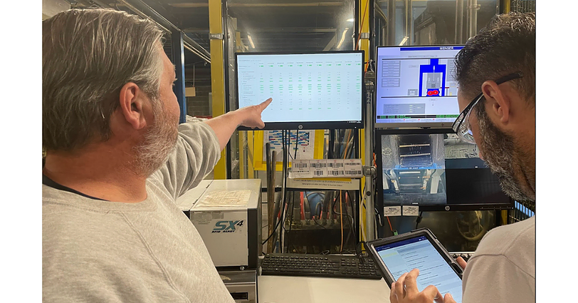 Two men are reviewing information on several screens in a control room. One man points to a large screen displaying detailed data, while the other holds a tablet. The room has industrial equipment and various technical displays.
