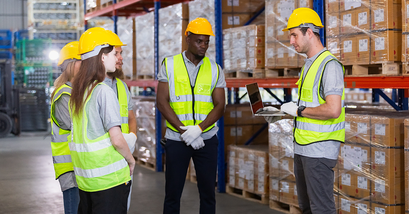 A supervisor in a safety vest and hard hat holds a laptop while talking to four warehouse workers. The workers, also wearing safety vests and hard hats, listen attentively. They are standing in an organized warehouse with shelves of boxed goods in the background.