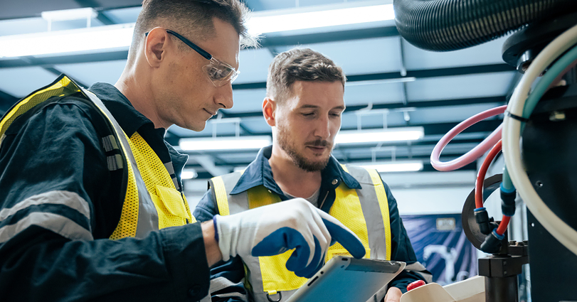 Two industrial workers, wearing safety glasses and yellow high-visibility vests, stand together in a factory setting. One worker is pointing to something on a tablet, while the other closely observes. They appear to be discussing or troubleshooting machinery.