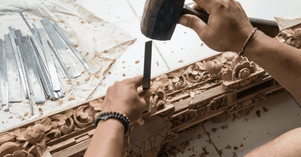 A person carves intricate designs into a wooden surface using a chisel and hammer. Metal chisels are scattered on a white cloth nearby. The detailed woodwork features floral and ornate patterns. The person's hands and tools are the main focus of the image.