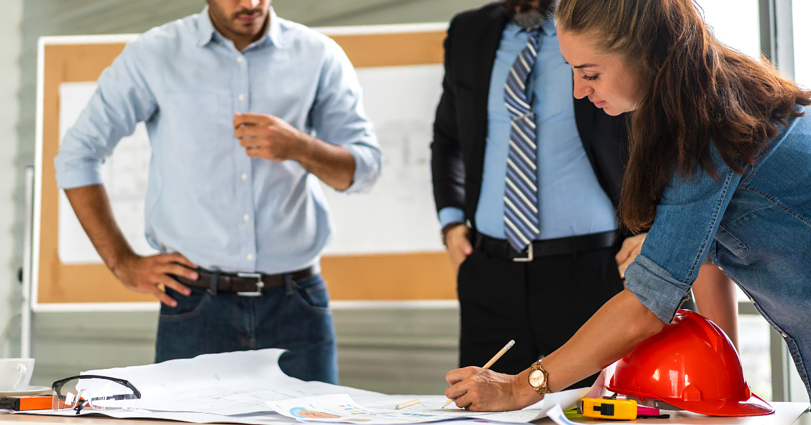 Three professionals in a meeting room discuss a project. A woman on the right writes on large sheets of paper, a red hard hat beside her. Two men, one in a suit and the other in casual attire, listen attentively. Blueprints and a level are on the table.