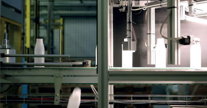An industrial production line with white bottles on a conveyor belt. The machinery appears to be automated, with metal frames and components. The background is slightly blurred, suggesting depth and a focus on the machinery and bottles in the foreground.