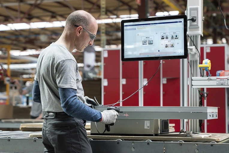 A bald man wearing safety glasses stands at a workbench in a factory. He is using a handheld device and looking at a large monitor displaying technical information. His work area includes red lockers and various industrial equipment in the background.