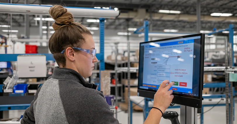 A person with long hair tied up and safety glasses is working in a factory setting, interacting with a touchscreen monitor displaying various data and options. The background features industrial equipment and shelving with electronic components.