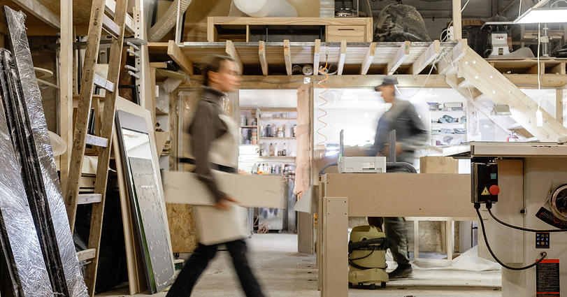 A woodshop with various tools, wooden boards, and shelving units. Two people are present; one is walking while holding a wooden plank, and the other works at a bench. The background features ladders, shelves, and a cluttered work environment.
