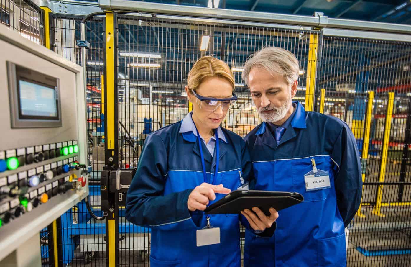 Two factory workers in blue uniforms stand in front of industrial equipment. The woman, wearing safety glasses, holds and points at a tablet while the man looks on attentively. Both wear name tags and are inside a facility with metal fencing and machinery.