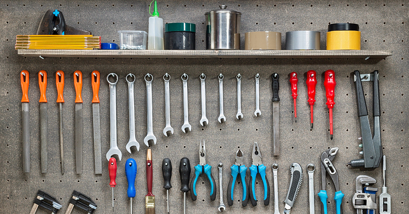 A neatly organized pegboard displaying various tools. On the shelf above, there are tapes, a brush, and containers. Hanging below are files, wrenches, pliers, screwdrivers, and other hand tools. The setup is orderly, with tools arranged by type and size.