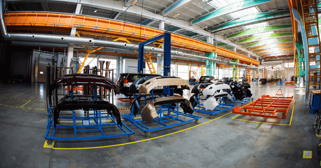 A spacious, modern factory floor featuring various car parts arranged on blue metal racks. Overhead cranes and other industrial equipment are visible, with bright lighting illuminating the high ceilings and clean, organized workspace.