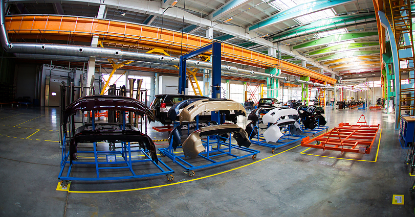 A spacious factory floor with a variety of car bumpers in different colors neatly arranged on blue metal racks. The background includes machinery, equipment, and overhead cranes, all under a high ceiling with bright lighting.