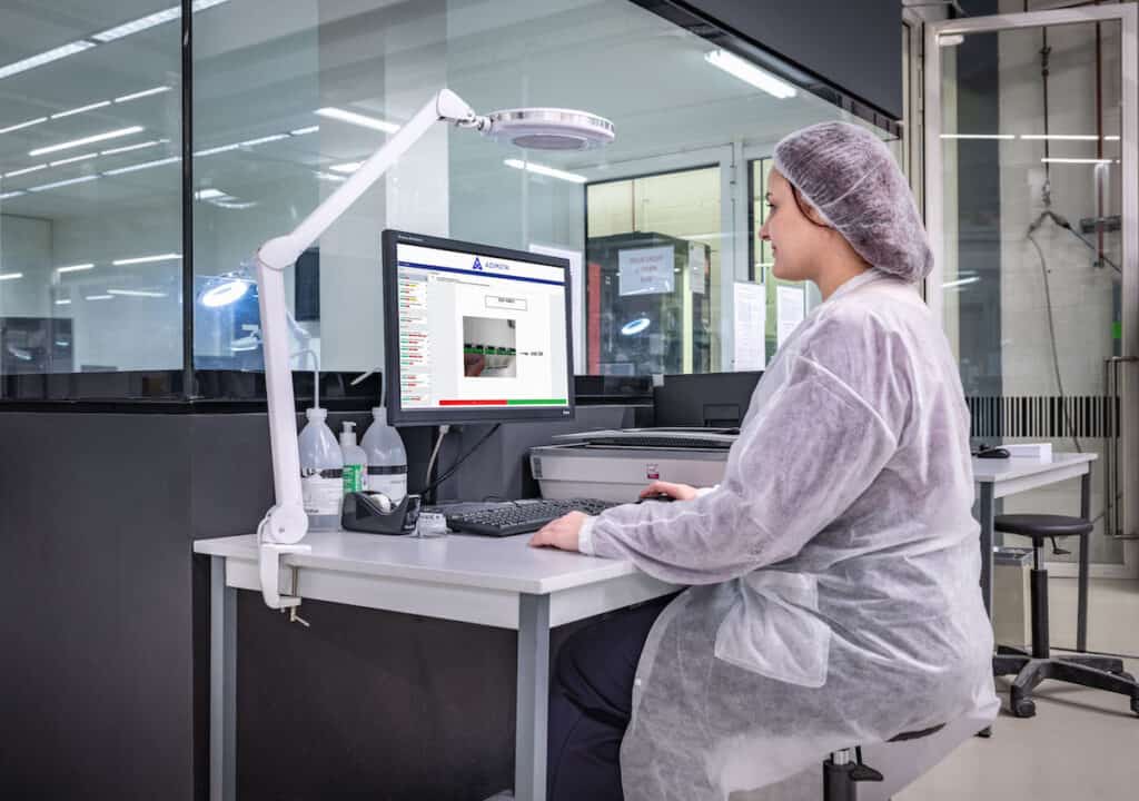 A person wearing a hair net and a lab coat sits at a desk in a lab setting, looking at a computer screen. The workspace features modern equipment, including a keyboard, mouse, and mounted magnifying light. The room is well-lit with a clean, sterile environment.