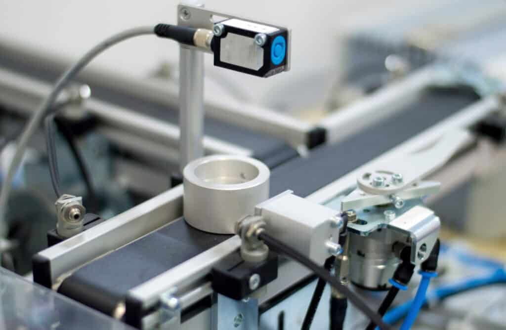 Close-up of an industrial machine featuring a conveyor belt moving cylindrical metal parts. The setup includes various sensors and mechanical components, with a focus sensor mounted above the conveyor. The machinery comprises metal and plastic parts with connecting wires.