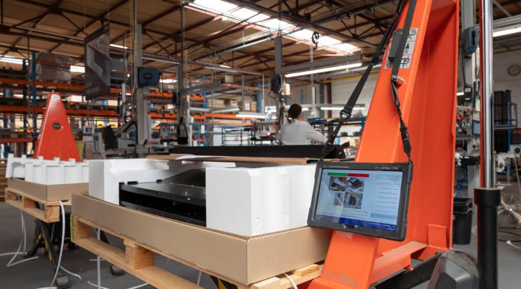 A factory floor with various machinery and products being assembled. In the foreground, there is a tablet mounted on an orange structure, displaying technical information. Workers are visible in the background, focusing on their tasks.