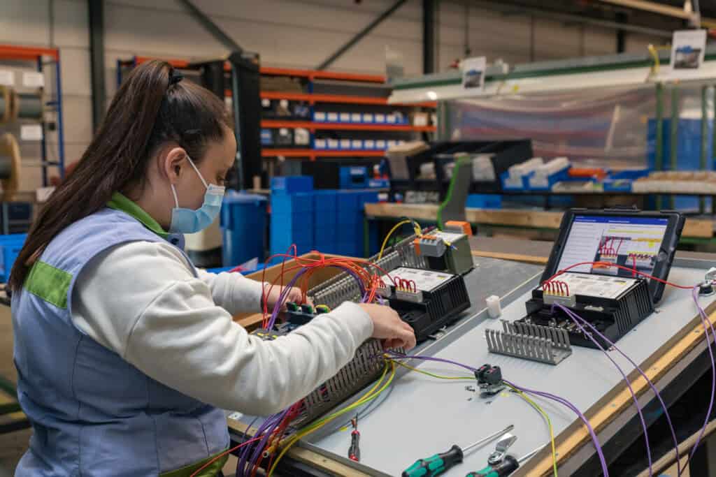 A woman wearing a face mask and protective vest works at a workstation with various cables and electronic components in a warehouse setting. Shelving units with organized bins and tools are visible in the background.