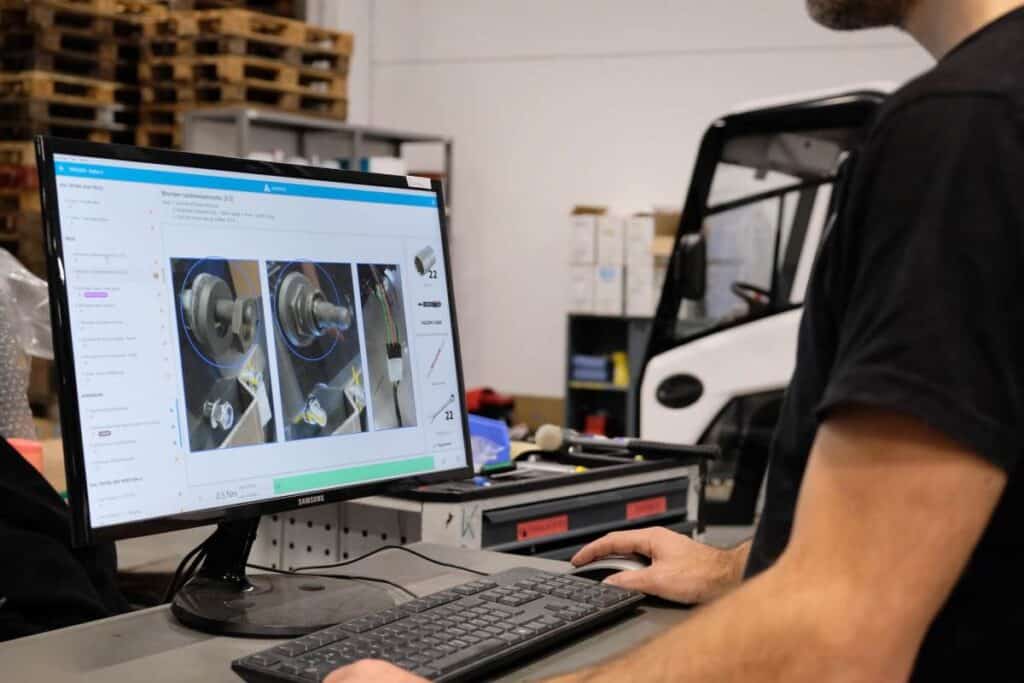 A person operating a computer in a workshop setting. The monitor displays a series of images of machine parts and diagrams. Shelves with various tools and equipment, including a white vehicle, are visible in the background.