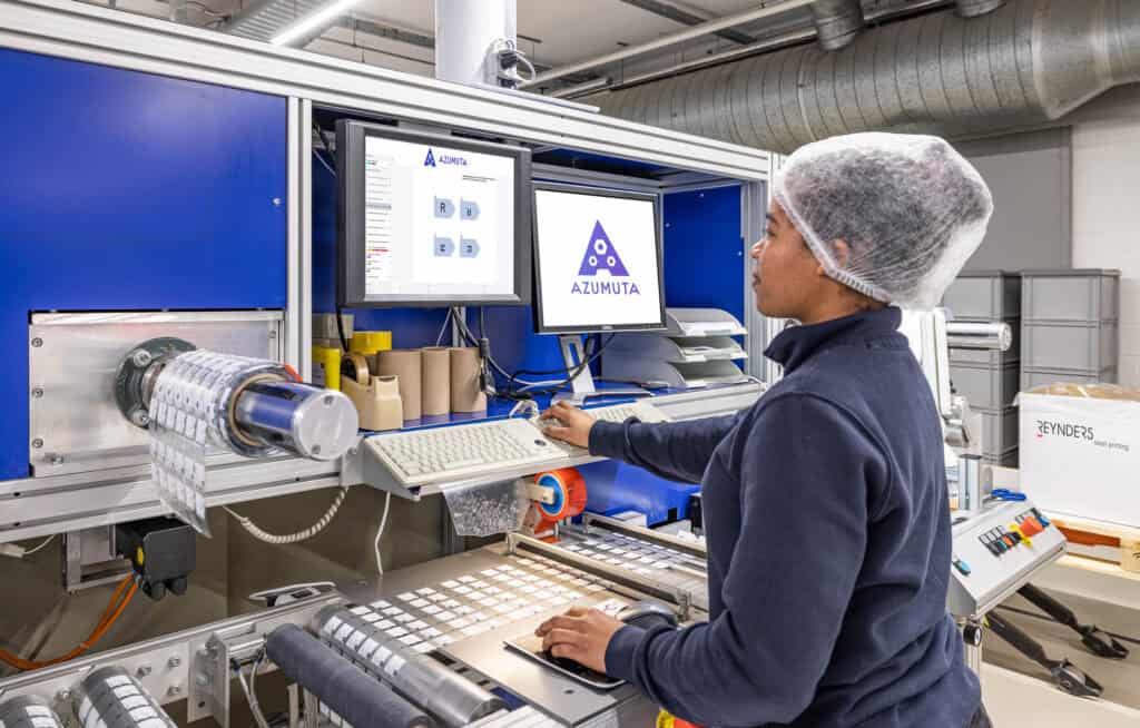 A factory worker wearing a hairnet operates a machine with a computer screen displaying the word 