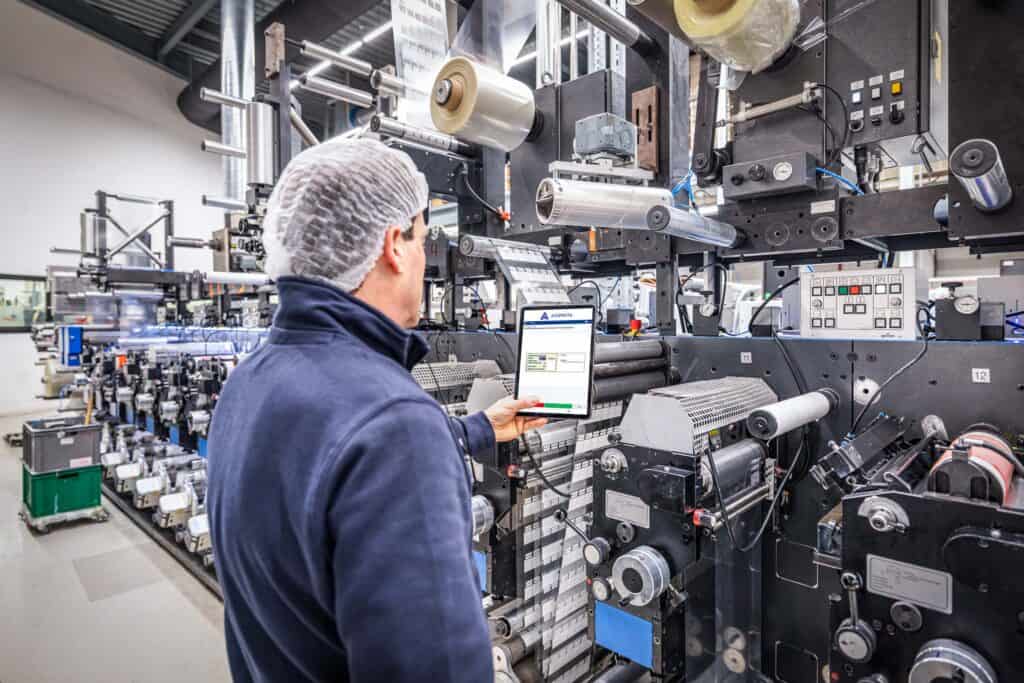 A person wearing a hairnet, glasses, and a blue jacket operates a large industrial printing machine. The person is using a tablet to control or monitor the machine. Various rolls of material and control panels with buttons and dials are visible.