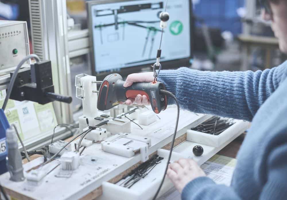 A person wearing a blue sweater is using a handheld scanner at a workstation equipped with various electronic components. In the background, a computer monitor displays a diagram with a green check mark.