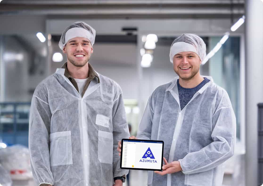 Two men wearing white lab coats, hairnets, and smiling, stand in a bright industrial setting. One holds a tablet displaying the Azumuta logo with a blue triangle. The background features machinery and blurred industrial equipment.