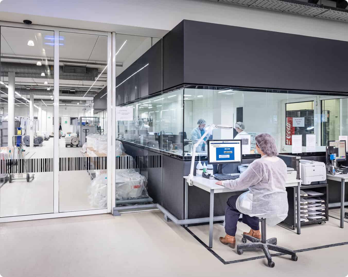 A laboratory worker sits at a desk with computer equipment inside a modern lab. The lab has glass partitions, and several other workers are seen inside a separate section working at lab benches. The environment is clean and organized with white and black decor.