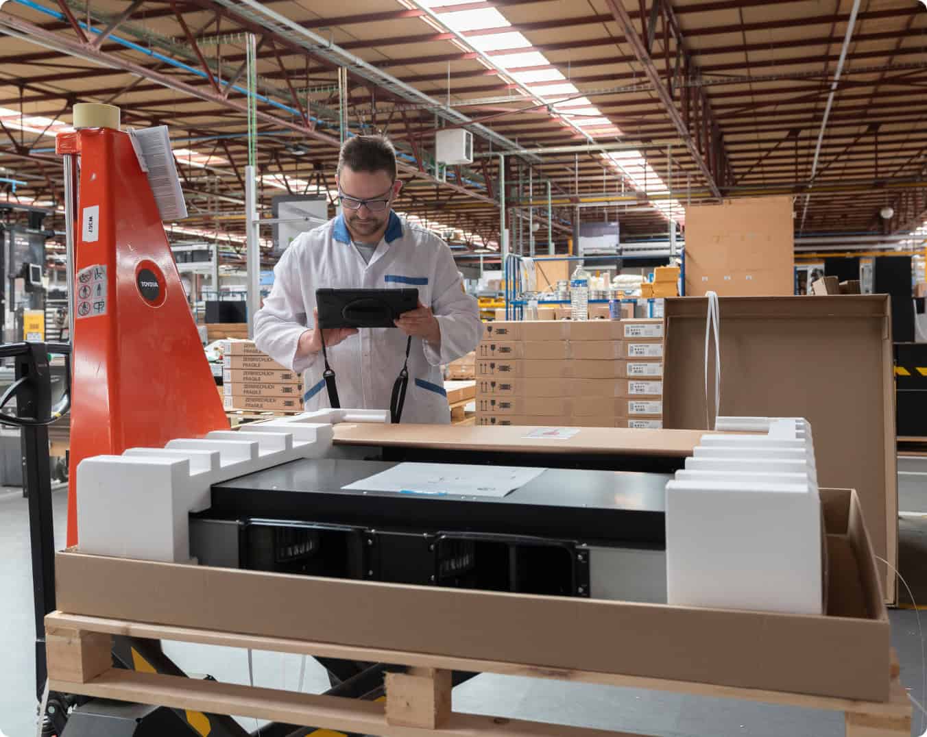 A man wearing a white coat and glasses stands in a factory or warehouse, holding a tablet. He is inspecting a large packaged item on a pallet, surrounded by stacks of boxes and industrial equipment under a high ceiling with skylights.