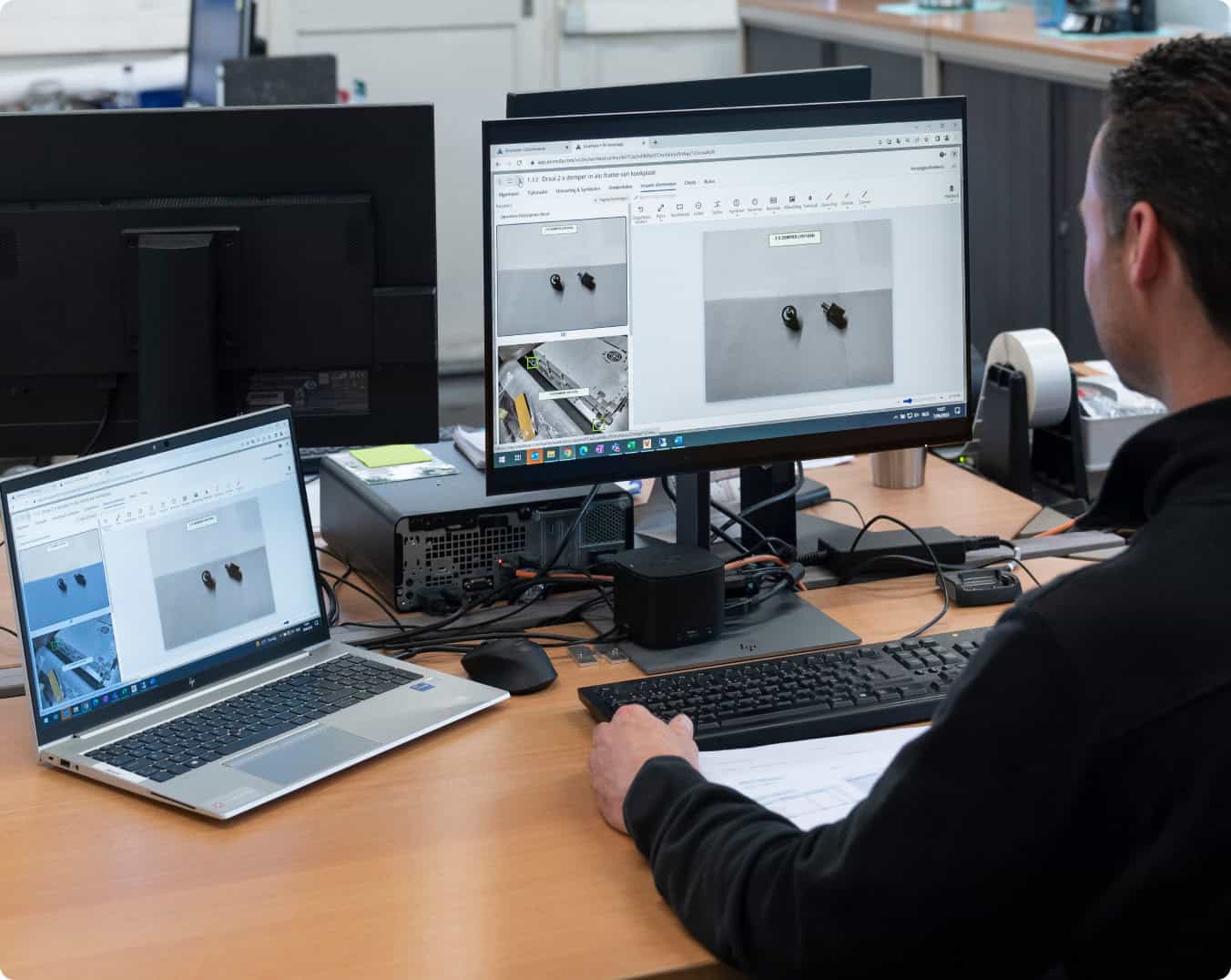 A person is working at a desk with a laptop and a desktop computer. Both screens display the same image of two small objects. The workspace includes various office equipment and documents are scattered on the desk. The person appears focused on their task.