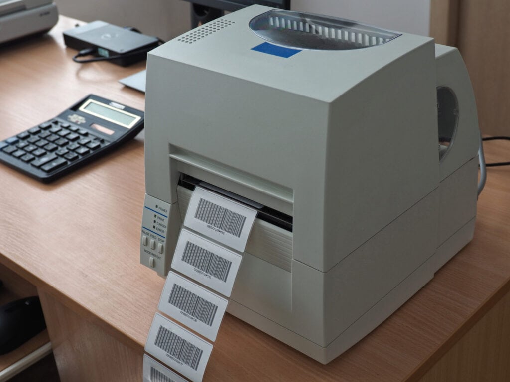 A barcode label printer on a wooden desk is printing a strip of barcode labels. Next to the printer, there is a black calculator and other office supplies. The setting appears to be an office or workspace.