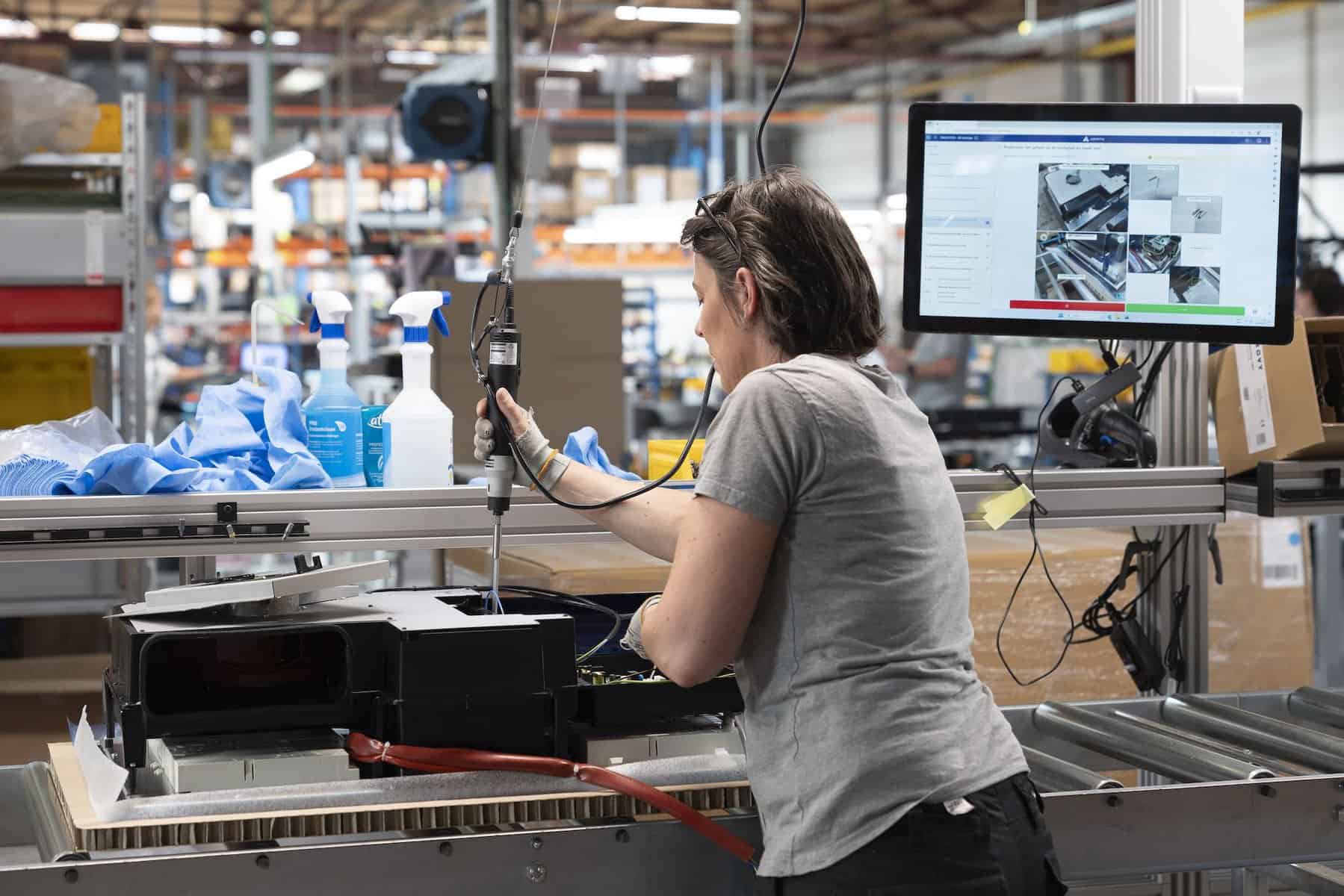 A person with short hair, wearing a gray shirt, is using a power tool at a workstation in what appears to be an assembly or manufacturing facility. In the background, there are shelves with supplies, cleaning products, and a computer monitor displaying instructions.
