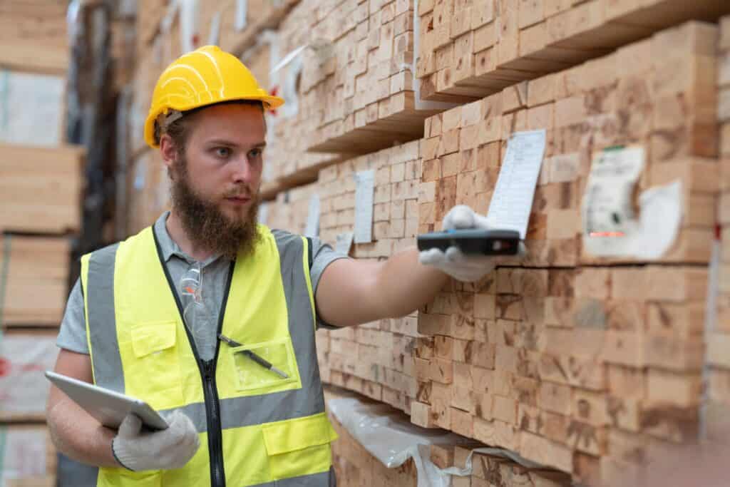 A man wearing a yellow safety vest and hard hat examines a stack of lumber in a warehouse. He holds a tablet in one hand and uses a device to scan a barcode with the other hand.