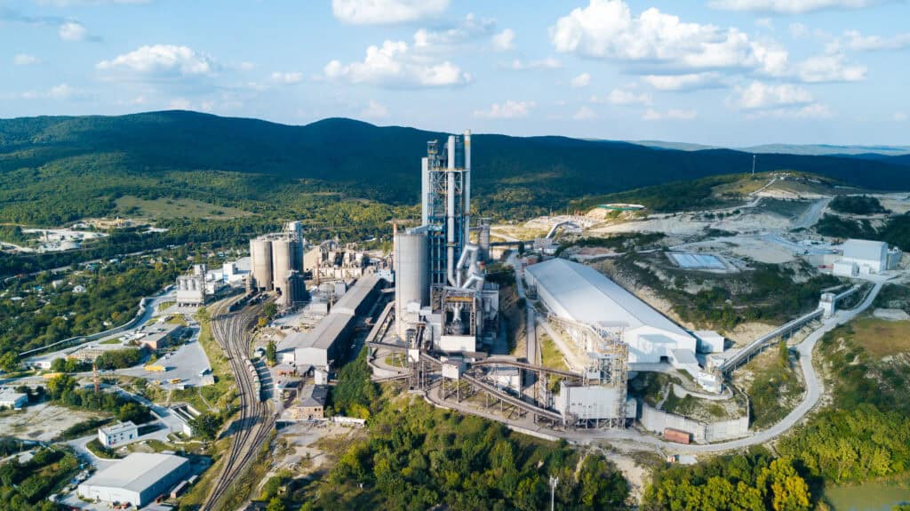 Aerial view of an expansive industrial cement plant surrounded by greenery and hills. The complex features tall chimneys, multiple structures, and winding roads connecting the various parts of the facility. The sky is clear with scattered clouds.