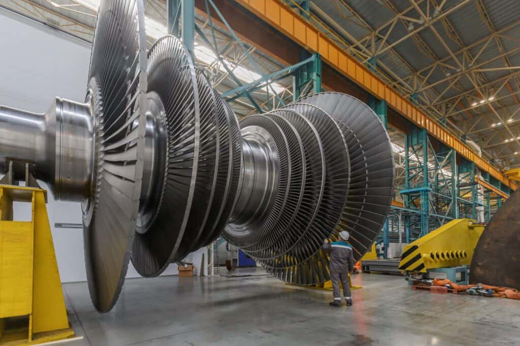 A large industrial turbine is displayed in a spacious factory setting with high ceilings and steel beams. A person in industrial work attire stands near the turbine, highlighting its massive size. Various equipment and tools are visible in the background.