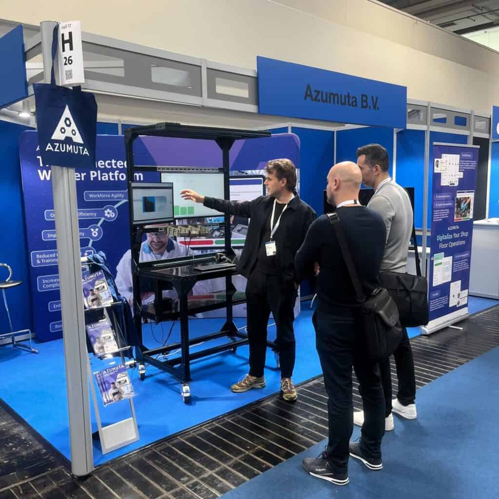 Three people stand in front of a booth at a trade show, where a man is presenting information on a monitor. The background includes banners displaying 