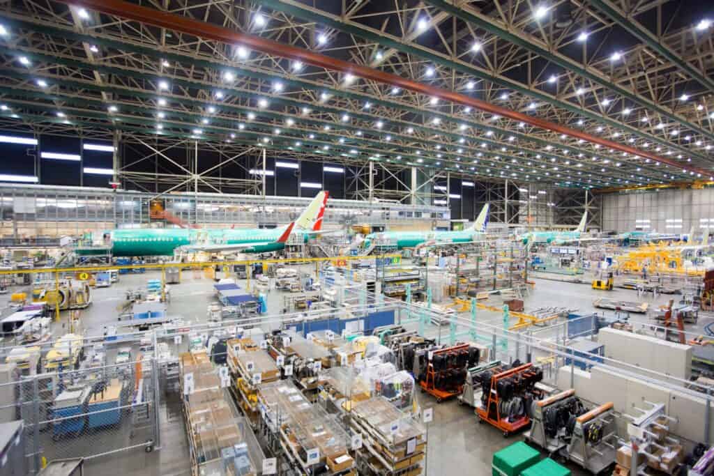 A vast airplane assembly factory with multiple aircraft under construction. The space is filled with machinery, tools, and parts. Bright lighting illuminates the large, open area, showcasing the complexity and scale of the manufacturing process.