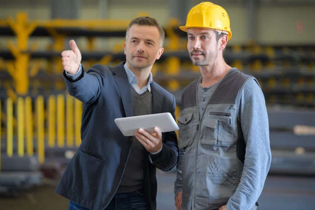 A man in a suit holding a tablet points forward while speaking to another man wearing a yellow hard hat and work vest in an industrial setting. The scene suggests a discussion or inspection related to the workplace.