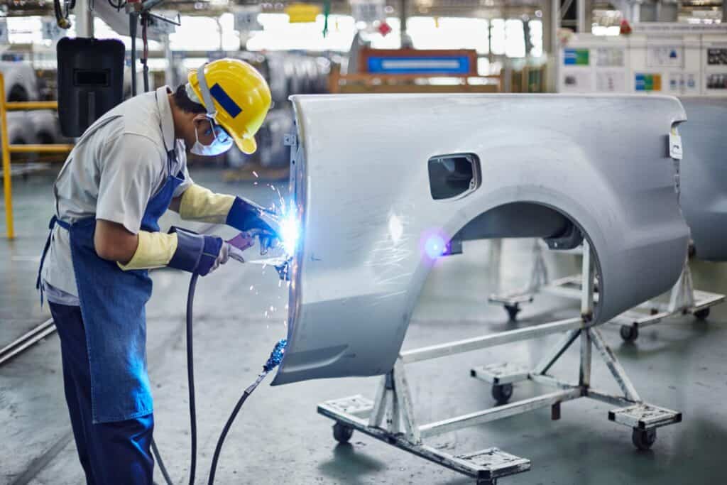 A factory worker wearing a yellow hard hat, safety goggles, and protective gloves uses a welding tool on a car door panel, creating sparks. The work area is well-lit with various machinery and equipment in the background.