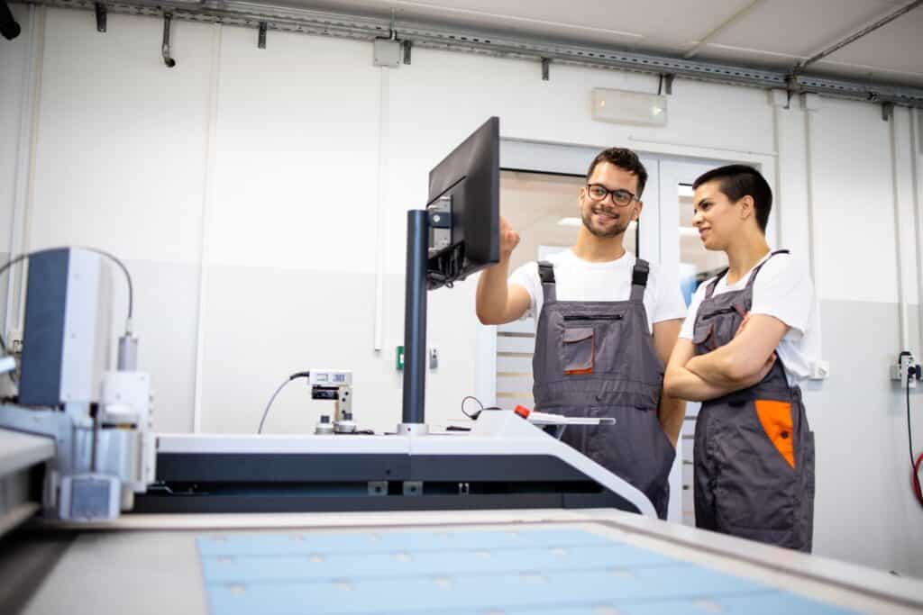 Two people in work uniforms stand in a modern industrial setting, looking at a monitor. One person is pointing at the screen while explaining something to the other. In the foreground, a large piece of machinery with a flat surface is partially visible.