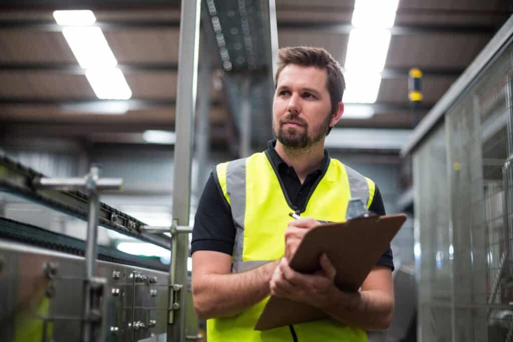 A man wearing a yellow safety vest and holding a clipboard stands in an industrial setting, likely a factory or warehouse. He appears focused and is looking upwards, possibly inspecting or taking notes about the surrounding equipment and machinery.