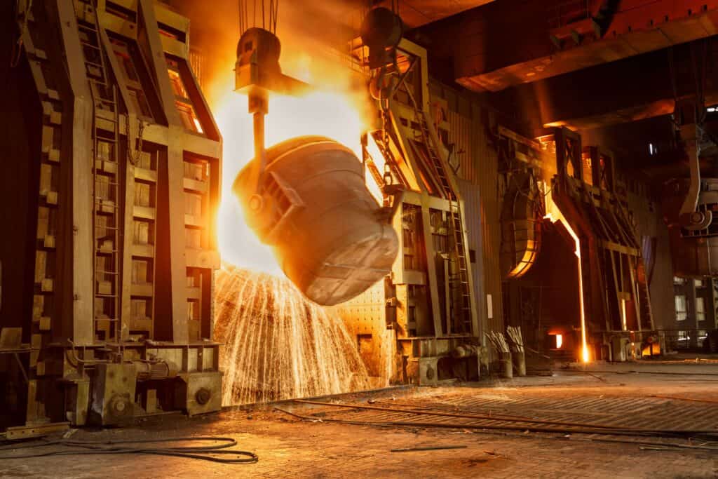 A large industrial facility with a bucket of molten metal being poured out, glowing brightly with sparks flying. The surrounding machinery and structures are illuminated by the intense heat and light from the metal.