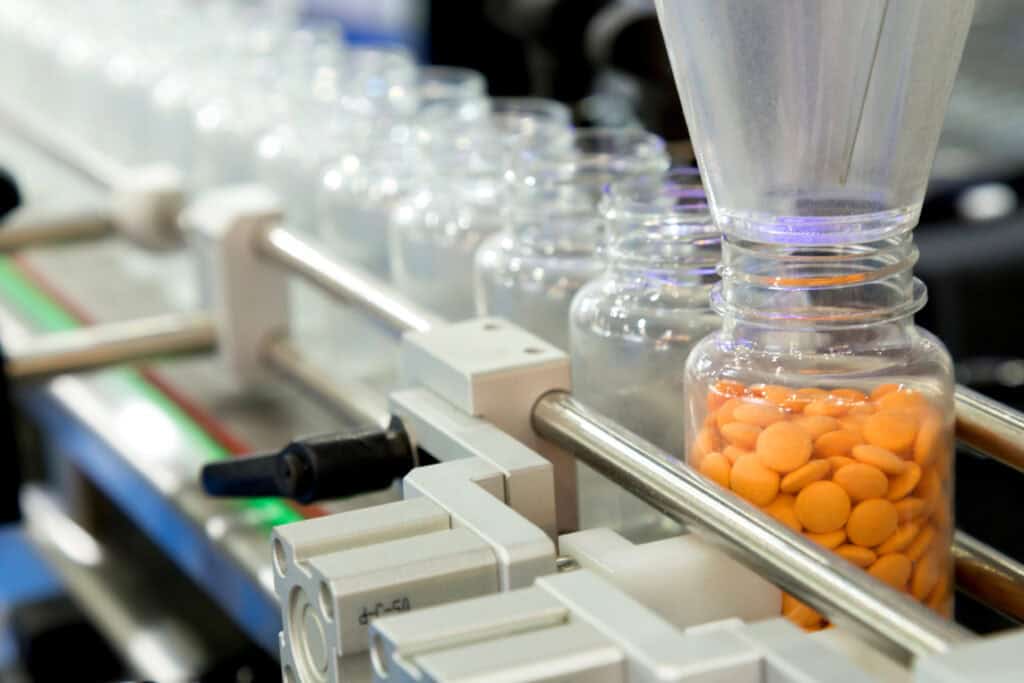 Pharmaceutical production line with clear bottles being filled with orange tablets by a dispensing machine. The bottles are aligned in a row on a conveyor belt, moving through the machine for automated filling. The background is blurred, emphasizing the process.