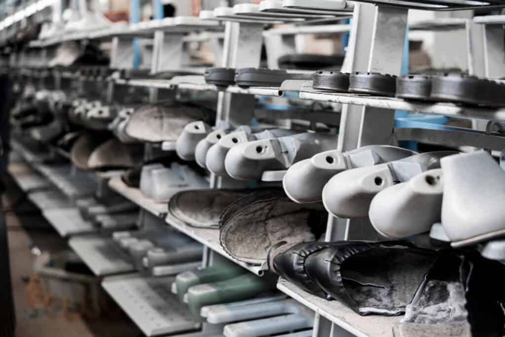 A row of leather shoes in various stages of assembly on a factory shelf. The shoes are placed on plastic shoe lasts, some without soles and others more complete, highlighting the process of shoe manufacturing.