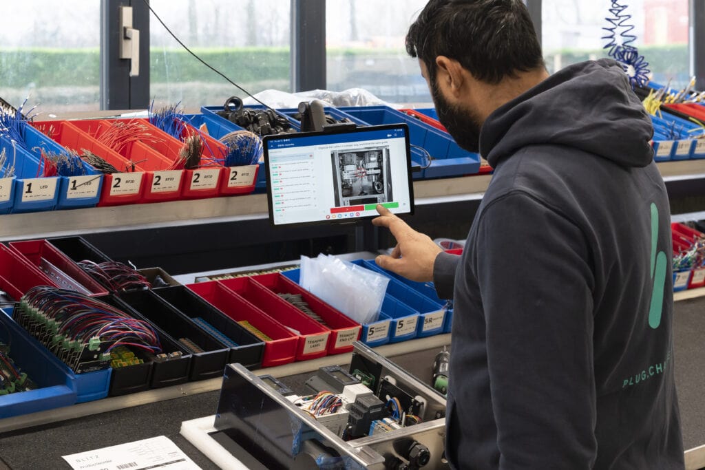 A man is working in an electronics workshop. He is interacting with a touchscreen monitor displaying technical diagrams. The workspace is organized with various colored bins containing components and tools. The man is wearing a dark hoodie with
