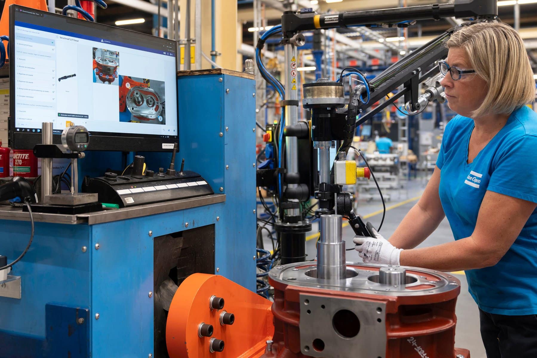 A female employee at a manufacturing plant is working with machinery while following digital work instructions displayed on a screen. The screen shows step-by-step visual instructions, including images of mechanical parts. The manufacturing environment has equipment and tools in the background, with the worker wearing protective glasses and a branded shirt, indicating a professional, organized workspace.