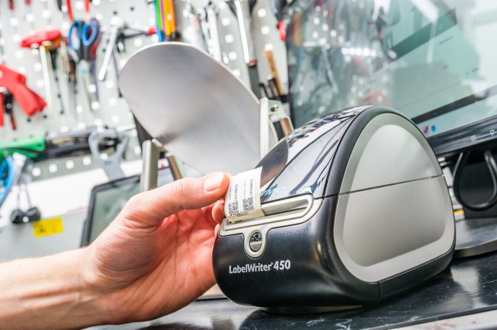 A close-up of a Dymo LabelWriter 450 printing a small QR code label in a workshop. A person’s hand is pulling the printed label from the machine. The background includes a workstation with tools, a computer screen, and other equipment, highlighting the use of printed QR codes for organization, labeling, or manufacturing processes.