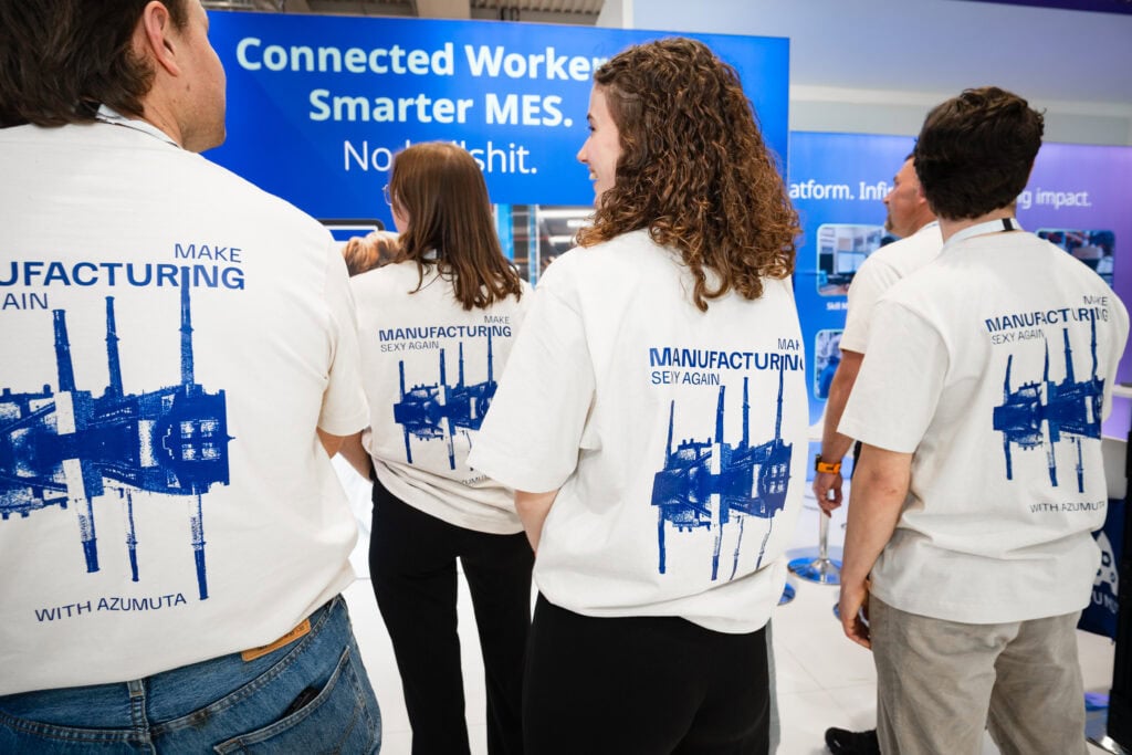 Azumuta team members at Hannover Messe 2025 wearing white T-shirts with the slogan “Make Manufacturing Sexy Again” and a stylized factory graphic. Booth backdrop with “Connected Workers. Smarter MES. No bullshit.” visible in the background.