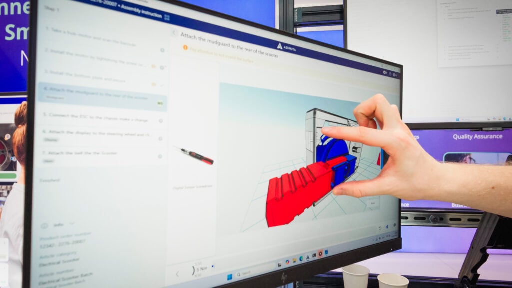 Close-up of a computer screen at the Azumuta booth showing a 3D digital work instruction for assembling a scooter. A hand gestures toward the screen, demonstrating interactive guidance in a real production scenario.